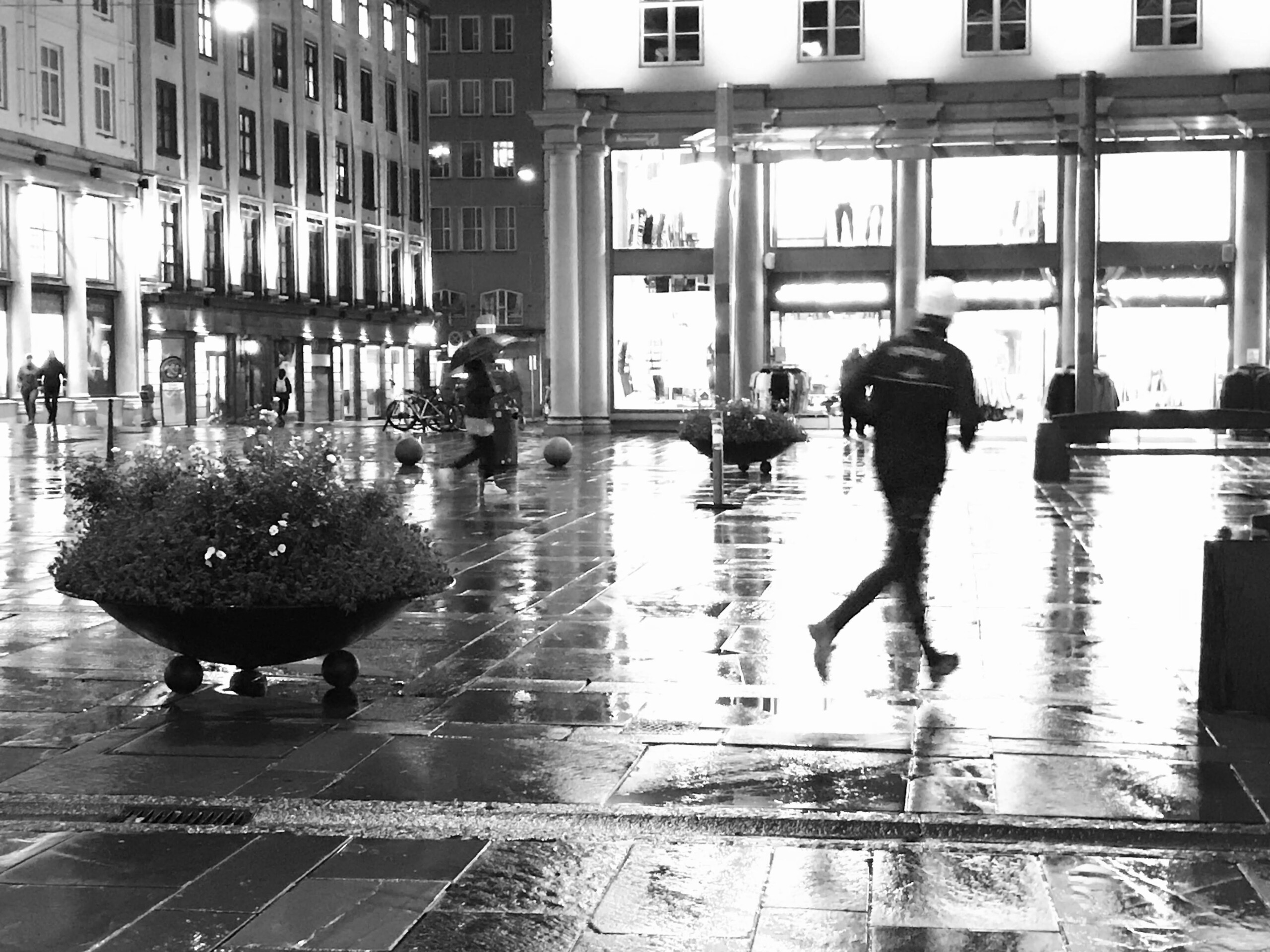 Svart-hvitt gatefoto av mennesker som beveger seg gjennom en regnvåt plass i Bergen om kvelden.
