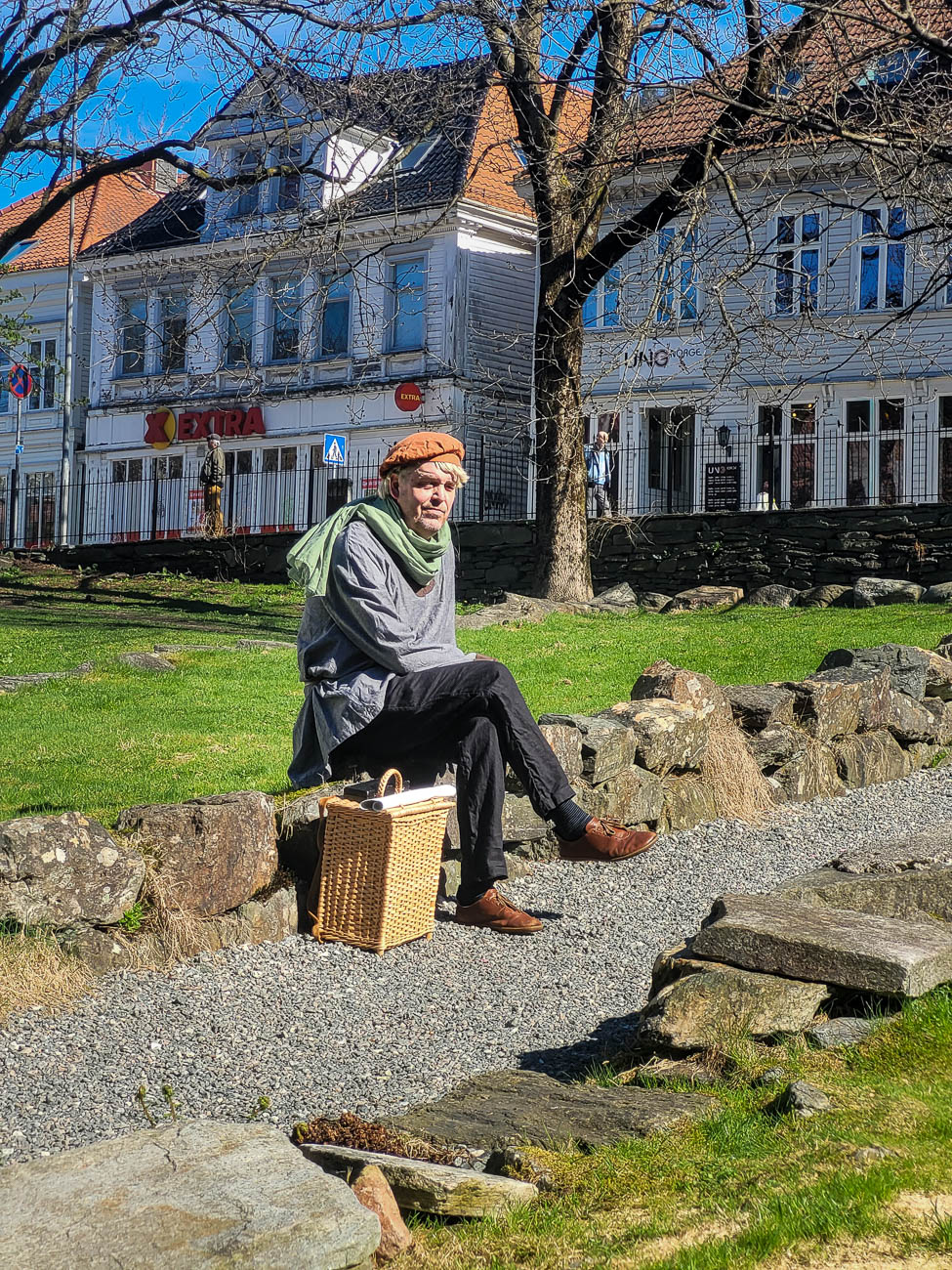 Person i historisk klesdrakt sitter ved Bryggen i Bergen med gamle trehus, grønt gress og sollys i bakgrunnen.